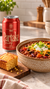A can of Partake Brewing Red non-alcoholic craft beer is shown next to a bowl of chili topped with sour cream, green onions, and cheese, with a slice of cornbread on a cutting board in a kitchen setting.